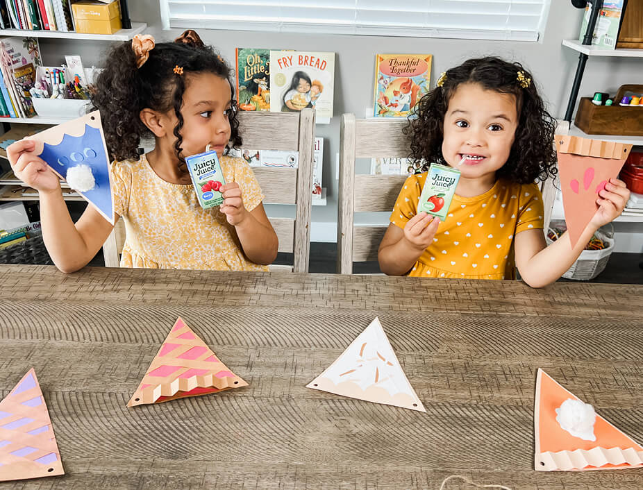 two young girls drinking juice juice and holding up their construction paper pies after they have been glued